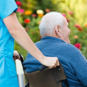 En kvinnelig omsorgsarbeider som triller rullestolen til en eldre mann med blomster i bakgrunnen. 